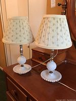 Two matching milk glass table lamps placed side by side on a wooden dresser, each with white pleated floral lampshades.