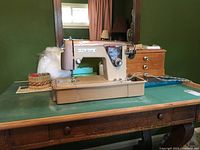Photo showing the Fleetwood portable sewing machine placed on a green cutting mat on a wooden table, with some sewing accessories including a spool of thread and a wicker basket.