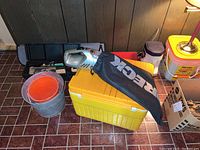 Wide view of buckets, cooler, Oreck car vacuum, hand tools, windshield sun screen, and cardboard box items arranged on tiled floor.