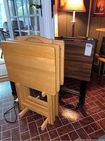 Two wooden TV tray stands: a blonde wood stand with three trays and a dark wood stand with two trays, placed on a tiled floor near a lamp and window.