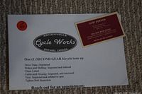 Photo of the $100 gift card paper voucher for a bicycle tuneup from Cycle Works and the business card of Kurt Dobson with contact details.