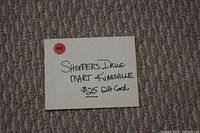Photo of a paper holder with handwritten label 'Shoppers Drug Mart Kingsville $25 Gift Card' on beige carpet background.