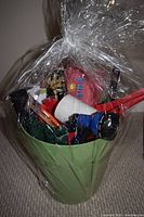Photo showing a green plastic planter pot wrapped in clear cellophane containing various gardening tools such as a red and black watering nozzle, white twine spool, orange handled shears, and other gardening accessories.