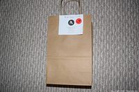 Brown paper gift bag containing the $50 gift certificate from Anna’s Flowers, with a white label attached showing the value.