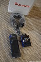 Photo showing black over-ear headphones wrapped in plastic along with a rectangular black headphone box and a boxed gaming mousepad with a galaxy design, all laid on a carpeted floor with a white The Source branded bag in background.