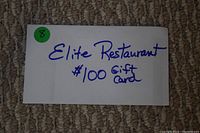 Photo of white envelope labeled "Elite Restaurant $100 Gift Card" in blue handwriting, placed on carpet.
