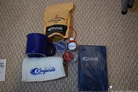 Photo showing items on carpet: coffee bag, blue mug, white toque, hand sanitizer, folded navy towel, and two small round bike lights in red and blue.