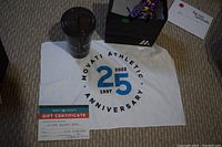 Flat lay photo of a white towel with Movati Athletic 25th Anniversary design, a 14 Day Pass certificate, and a black smoothie cup with lid on carpet.