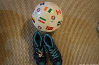 Photo of a white size 5 soccer ball decorated with multiple international flags and EURO 2016 logos, shown alongside two black and blue soccer shoes with turquoise laces.