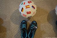 Photo showing a soccer ball decorated with international flags and a pair of black and blue soccer shoes labeled size 6.