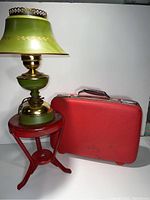 Red metal stool supporting a green and brass table lamp and next to a red American Tourister suitcase.