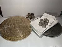 Full view showing 8 gold placements stacked, 8 silver placements stacked, 9 white cotton tea towels folded and 8 silver rhinestone napkin rings on top, plus 8 metallic silver textured napkin rings grouped behind.