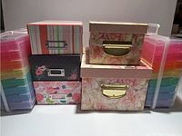 Front view of five decorative cardboard organizer boxes with colorful patterns and metal/gold label holders stacked in center. Two plastic multi-drawer craft organizers with rainbow drawers on each side.