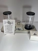 Twin Tornado Mixer base with two clear plastic blending containers, each with black lids, placed on a white surface, showing measurement markings and brand logo.