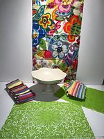 All items displayed: 4 green laser cut placemats, 8 colorful striped napkins, floral fabric table runner, cream ceramic fruit bowl on pedestal base.
