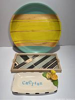 View of all three trays stacked: large round colorful wooden tray in back, rectangular wooden tray with geometric design in front, ceramic tray with flower detail and Cherish text at bottom.