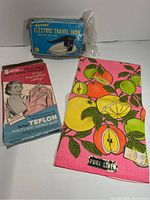 Photo of blue box labeled 'Tajpont Electric Travel Iron' with plastic packaging beside it, colorful linen tea towel with fruit print.