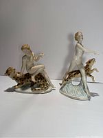 Photo shows front view of two vintage porcelain statues: one seated nude woman on a panther, one standing nude woman posing with a panther on a water-like base.