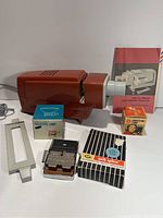 Photo showing red vintage projector with accompanying manual, boxed projector lamps, and HP quik splice splicer.