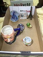 Full view of lot showing all six collectible items inside a cardboard box including a glass paperweight, two porcelain dolphins, ceramic bird egg cup, a painted face mug, and a metal Eiffel Tower model.