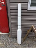 Full view of white painted solid wood column post standing upright against siding and next to a red vending machine, showing its full length and design.