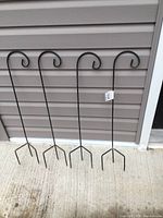 Four black metal garden hooks standing upright against a vinyl siding wall, showing the full length and curved hook on top.