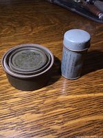 Photo showing the two military tins on wooden surface, one round and one cylindrical, displaying overall shape and wear.