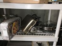Shelf with stacked aluminum grilling trays in plastic foil and assortment of pots, pans and lids including stainless steel and non-stick items