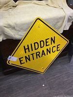 Photo showing front of the reflective yellow road sign with black text 'HIDDEN ENTRANCE' and mounting holes.