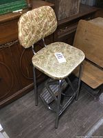 Photo showing vintage chrome framed folding step stool with patterned upholstered seat and back, fold out step visible, placed on a floor beside wooden furniture.