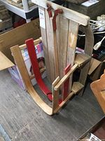 Photo showing child's wooden sleigh from an angled top view, featuring curved wooden runners partially painted red, folding handle with metal hinges and leather straps, and vertical wooden slats forming the seating surface.