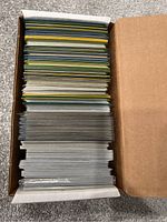 Top view of a cardboard box filled with a large number of loose Pokémon cards stacked vertically, showing card edges with different colors and some sleeved cards near the bottom.