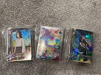 Three Naruto holographic trading cards in plastic sleeves on carpet: Itachi Uchiha, Tsunade, and Kakashi Hatake