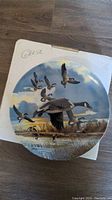 Round Dominion China collector plate showing Canada geese flying over a marsh, mounted on top of a white box with 'Geese' written on it.