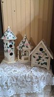 Photo of 3 decorative hand-painted wooden birdhouses in cream color with floral designs, displayed on lace cloth against wooden panel background.