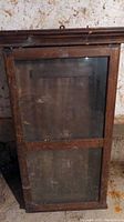 Front view of antique hanging display cabinet showing glass door and wooden frame with hanging hook on top.