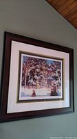 Framed print showing full window view with snow-covered trees, house, and two figures in winter clothing. Professionally framed in dark wood with cream mat and green border.