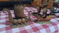 Two small metal ship figurines on a red and white checkered surface. One is a Viking longship, the other a multi-mast sailing ship.