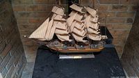Photo showing the full miniature sailboat model from the front, displayed on a black surface against a brick background. The ship has detailed sails and rigging.