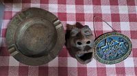 Photo showing all three items on a red and white checkered cloth: round metal ashtray, carved face ashtray, and no smoking sign