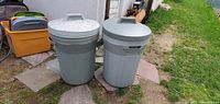 Two grey 39 gallon Sears garbage cans with lids placed on stone tiles outside near grass and other containers.