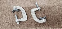 Two silver-colored metal C-clamps shown on carpeted surface, each with threaded screw and rubber clamping pads.