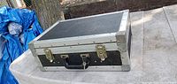 Closed view of the black and silver snap case showing metal edges, corners, latches, and handle on a table outdoors.