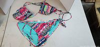 Photo showing the full bikini set with top and bottom laid out, displaying vibrant pink, blue, and multi-color geometric pattern and bead detailing on the top ties.