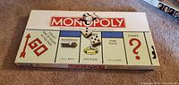 Classic Monopoly board game box top showing the game logo and iconic gameplay spaces like Boardwalk and Park Place, with dice and a token positioned.