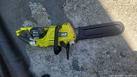 Full view of the Ryobi cordless chainsaw showing the entire tool laid down on concrete surface with protective bar cover in place.