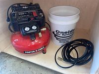 Full view of red Porter Cable 6-gallon air compressor with gauge panel, black motor housing, next to white Oneida Air Systems bucket and coiled black air hose on wooden shelf.