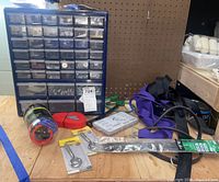 Hardware organizer unit with small drawers filled with screws and fasteners, placed on wooden surface alongside straps, hooks, zip ties and stretch cords.