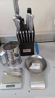 Photo showing kitchen knife block with multiple knives, stainless steel flour sifter, spoon rest with note, digital kitchen scale.