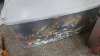 Clear plastic tote filled with various mixed Lego pieces on carpet floor.
